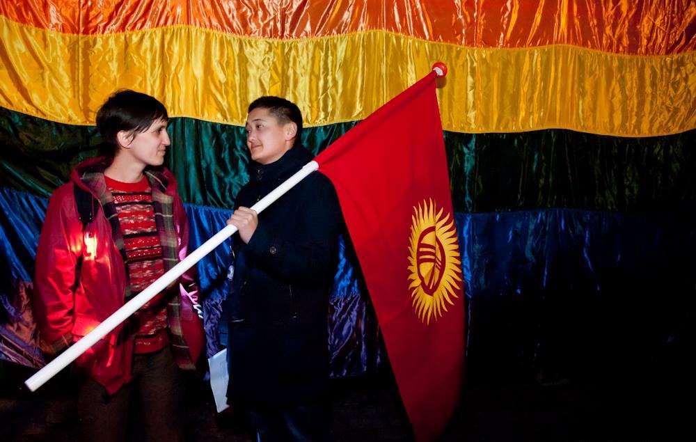 Изображение: Neeltje Knaap / Фото из личного архива героини материала, Назик. Два человека смотрят друг на друга, стоя на фоне радужного флага. На человеке слева красная куртка и свитер. На человеке справа черная одежда, в левой руке он_а держит флаг Кыргыстана.