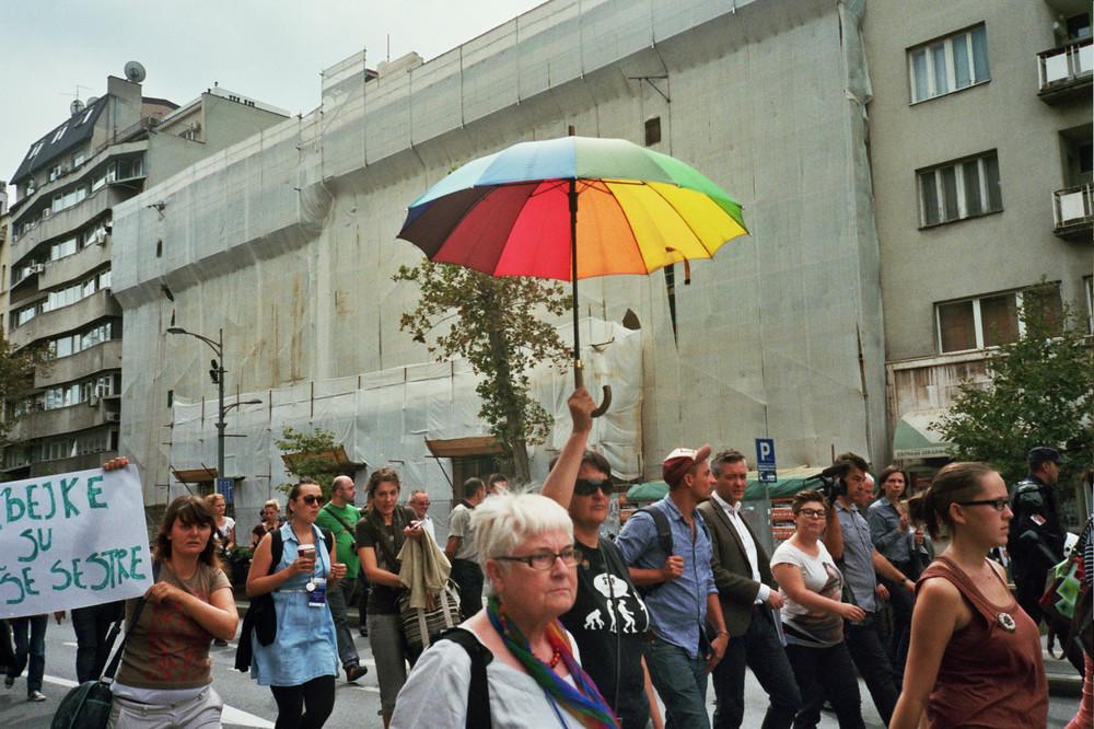 Изображение: Lazara Marinkovic / Лесбийский марш в Белграде, 2015 год. На фото: группа людей идёт по проезжей части вдоль большого серого здания. Человек по центру снимка высоко поднимает зонтик радужных цветов.